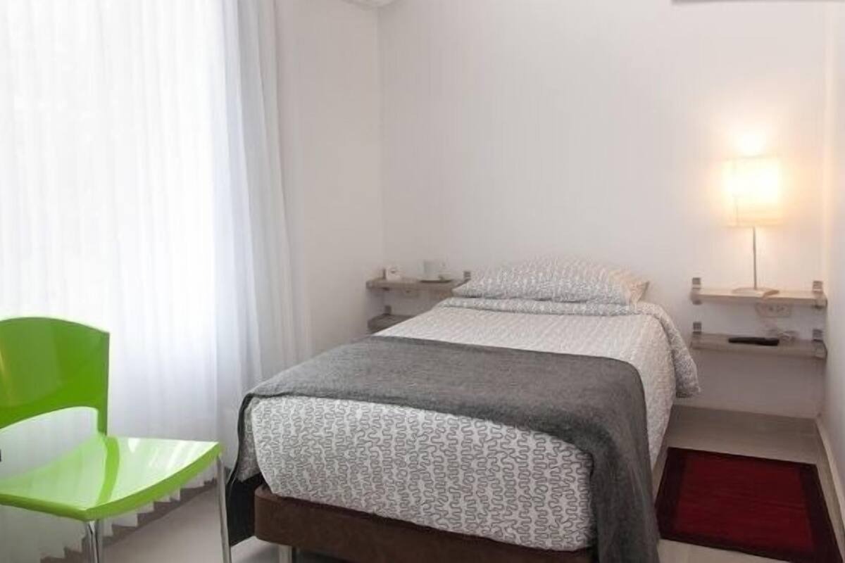 A small, clean room with a bed, a green chair, a bedside table, and a lamp.