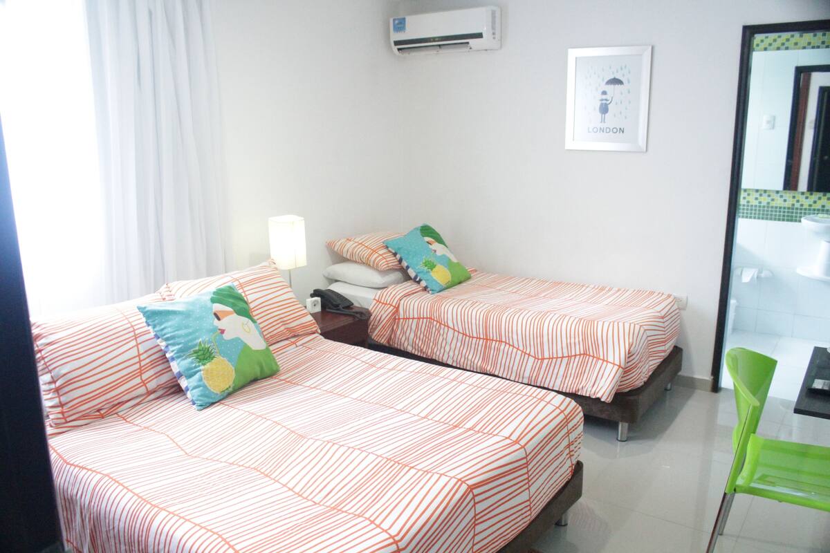 A hotel room with two beds, each with striped bedding and pillows, a bedside table with a lamp, a framed picture on the wall, and a green chair.
