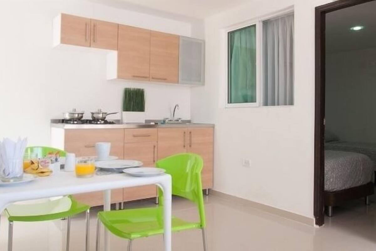 A compact kitchen with wooden cabinets, a dining table with chairs, and a bed in the adjacent room.
