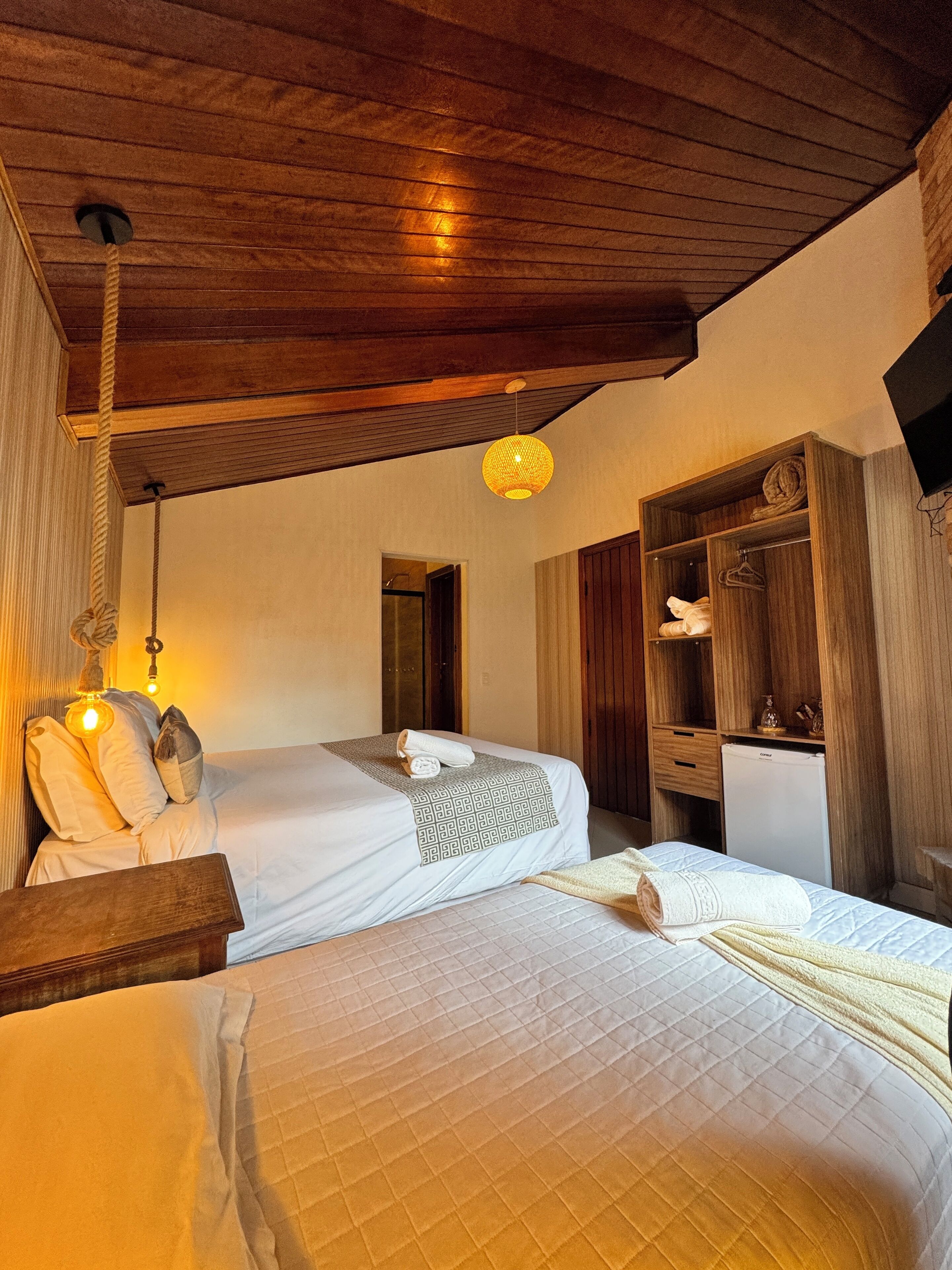 A hotel room with two beds, wooden ceiling beams, a wooden wardrobe, and a small white refrigerator.