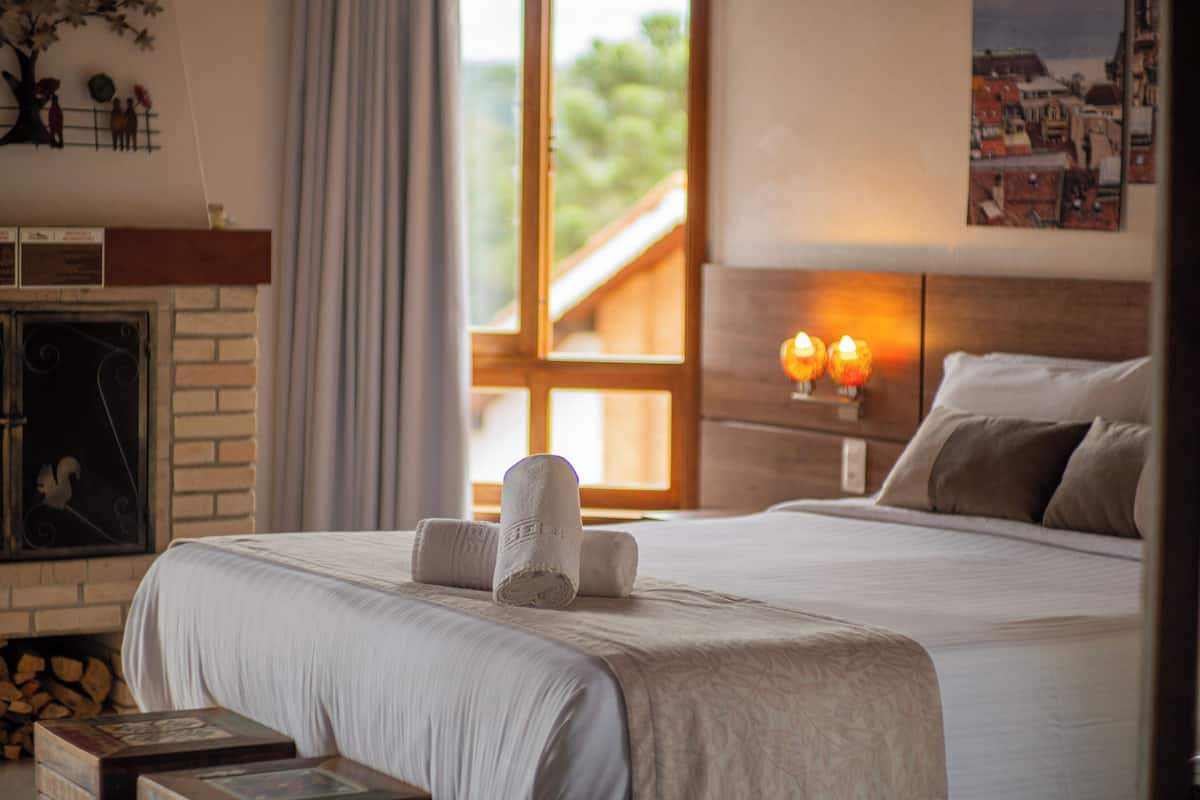 A neatly made bed with rolled towels, a fireplace, and a view of a wooden deck through a window.