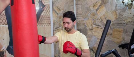 Una persona practica boxeo con un saco de boxeo rojo en una habitación con paredes de piedra y una cinta de correr al fondo.
