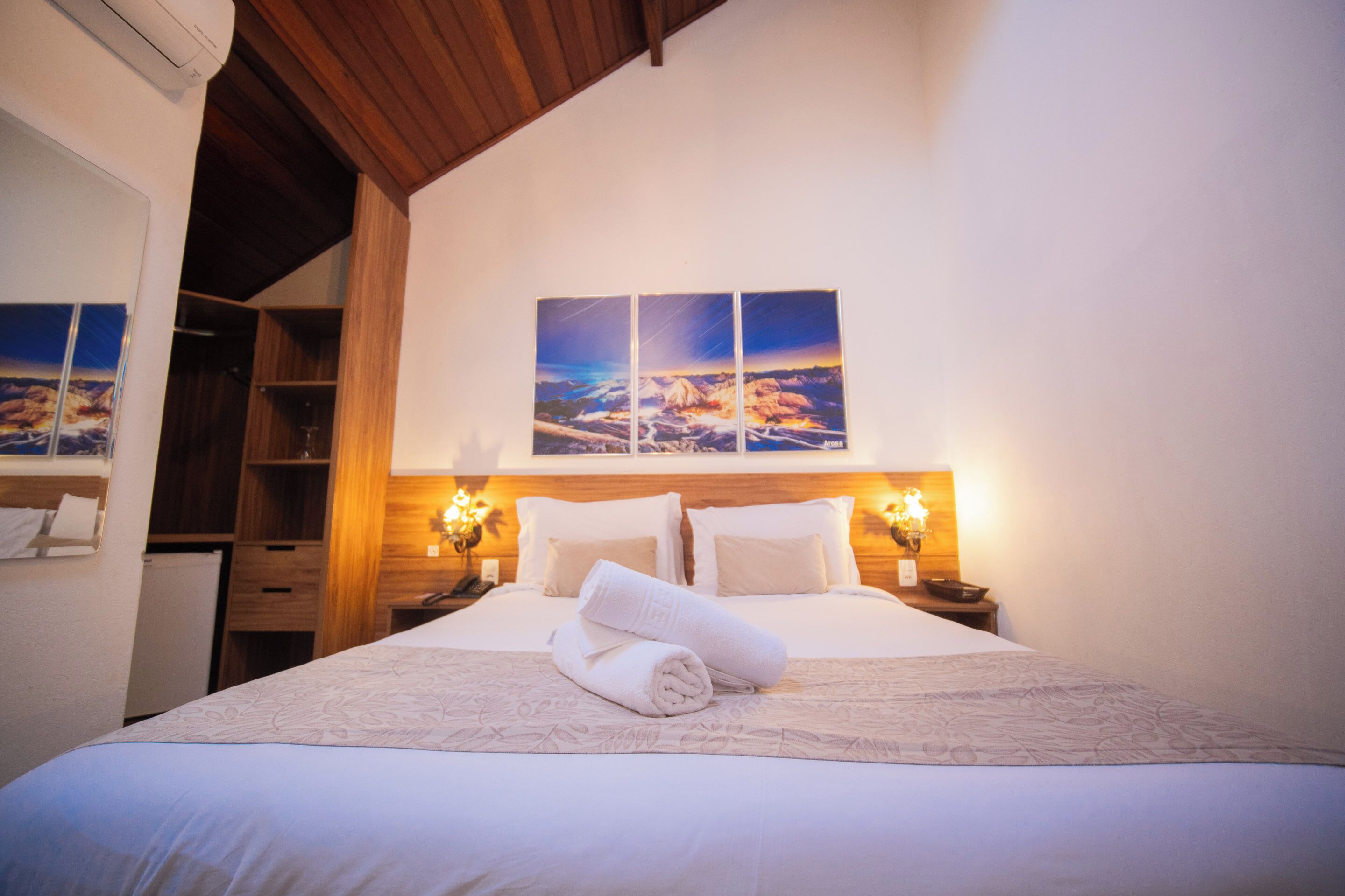 A neatly made bed with a rolled towel, wooden headboard, and a wall-mounted picture.