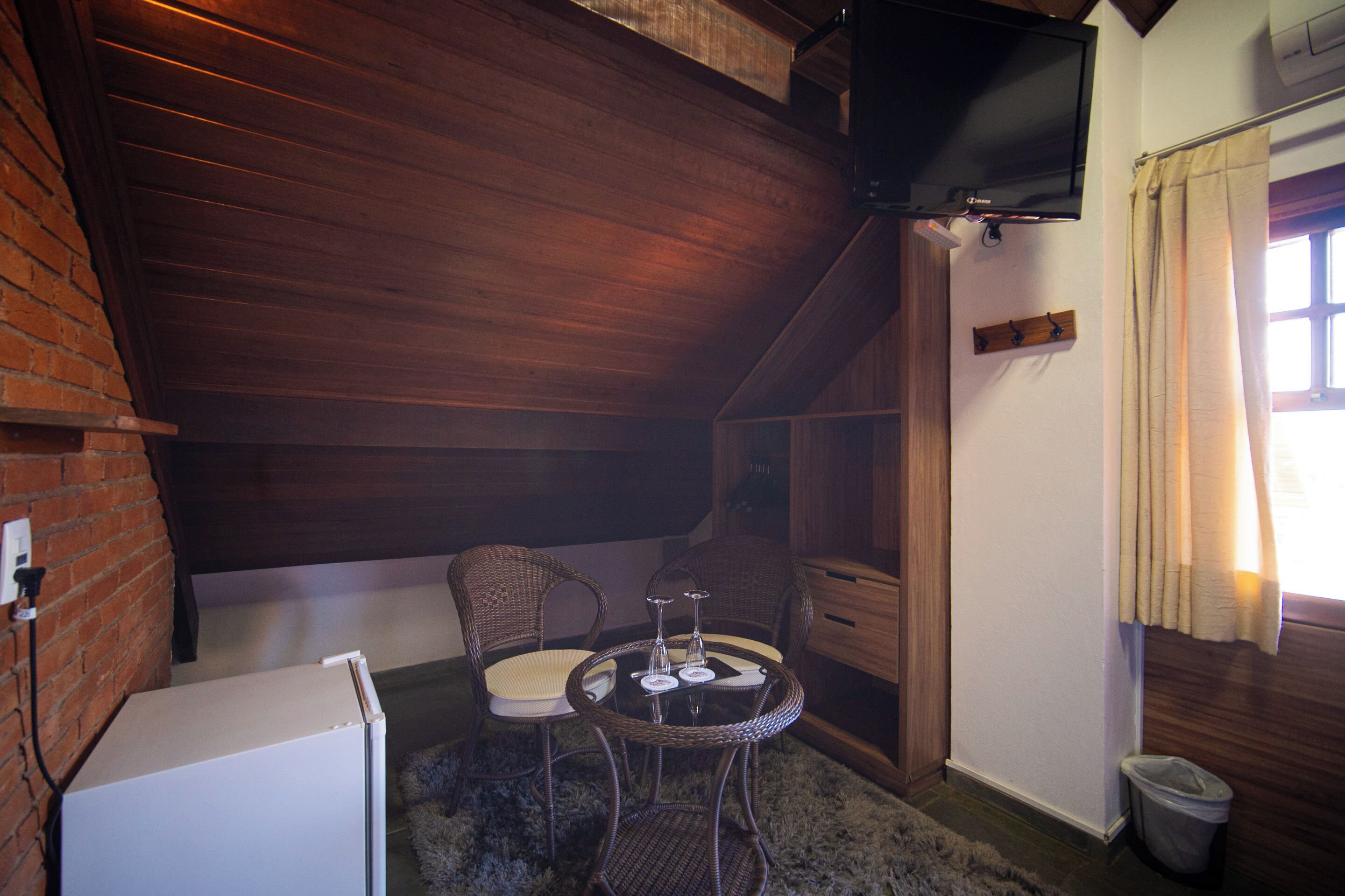 A cozy room with a brick wall, a television mounted on the ceiling, a small table with chairs, and a window with curtains.