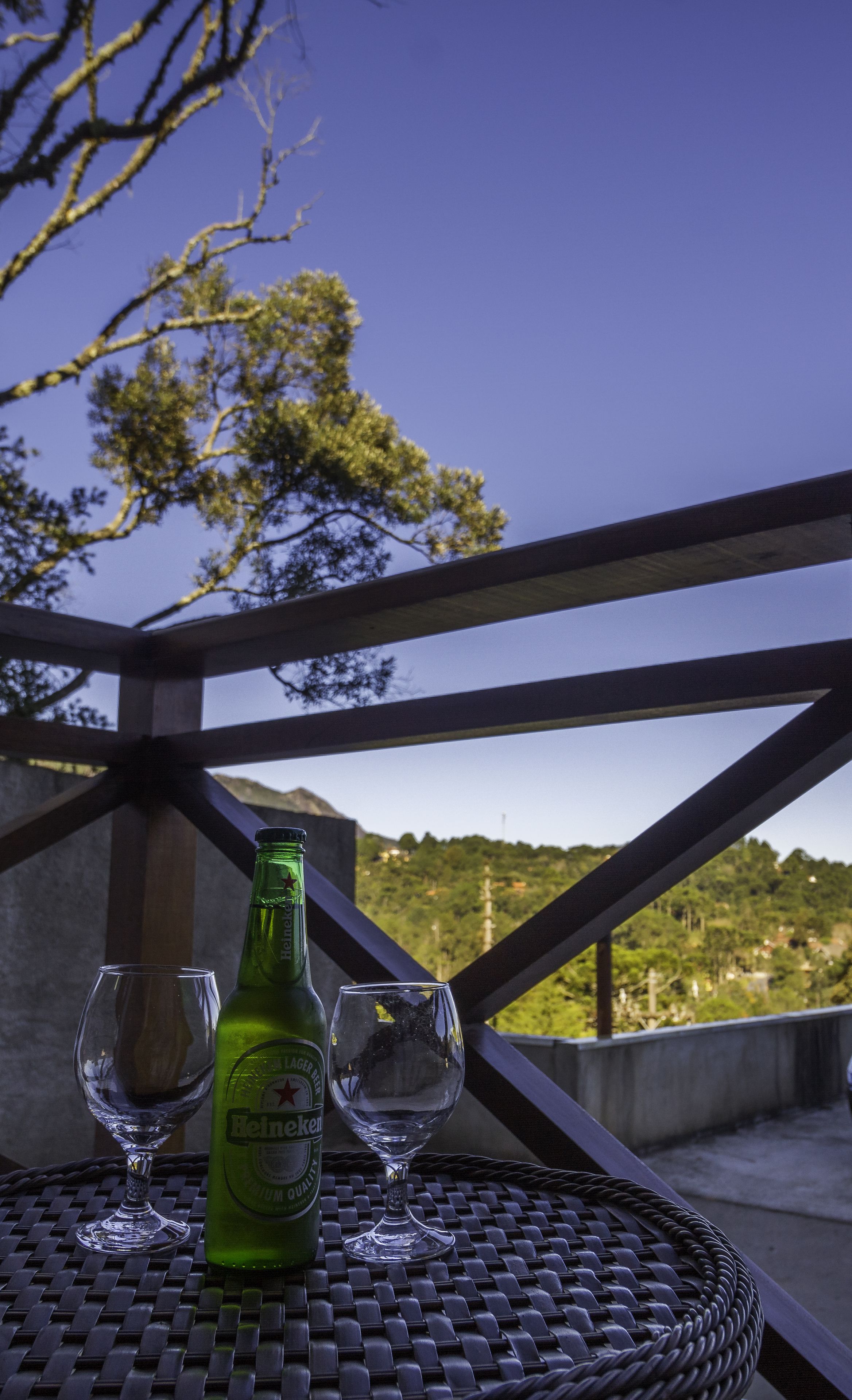 Een groene fles Heineken bier, twee lege glazen en een rieten tafeltje op een balkon met uitzicht op bomen en heuvels.