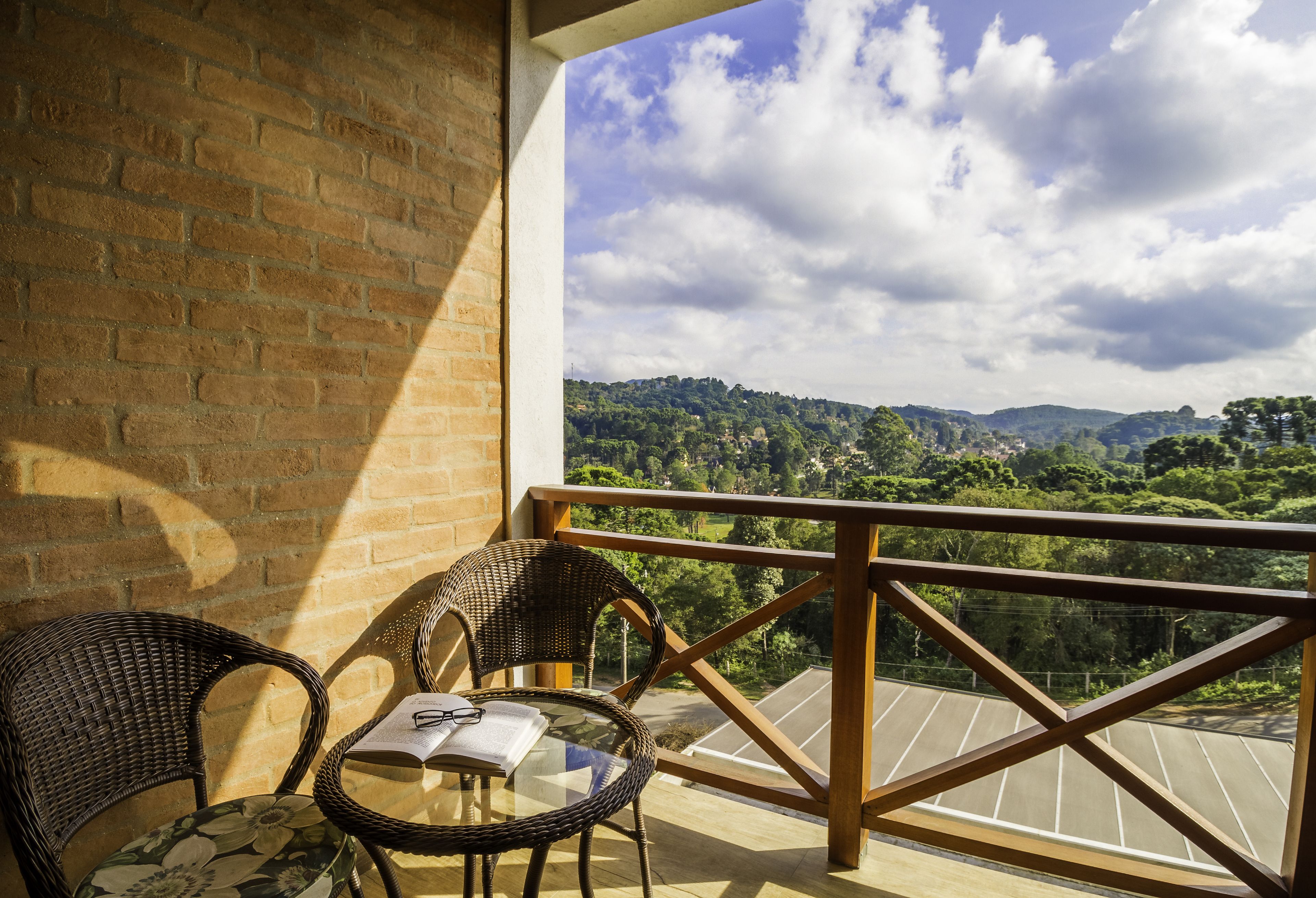 Uma varanda com cadeiras de vime e uma mesa, com vista para uma paisagem pitoresca com árvores e colinas.