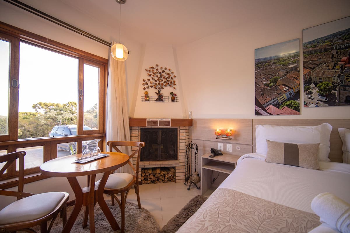 A hotel room with a fireplace, a dining table with chairs, a bed with a large window, and a cityscape painting on the wall.