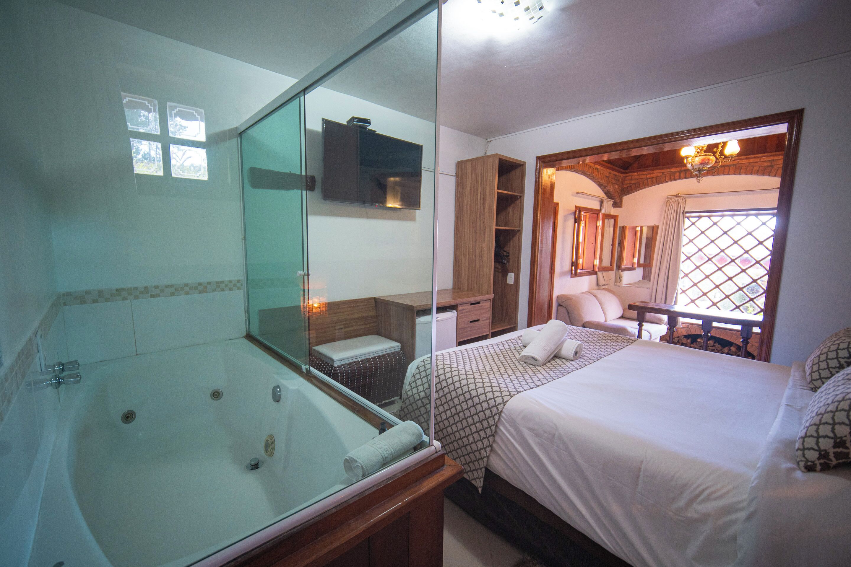 A hotel room with a glass-enclosed jacuzzi, a bed with patterned bedding, a wooden ceiling, and a view of the outdoors through a window.