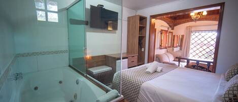 A hotel room with a glass-enclosed jacuzzi, a bed with patterned bedding, a wooden ceiling, and a view of the outdoors through a window.