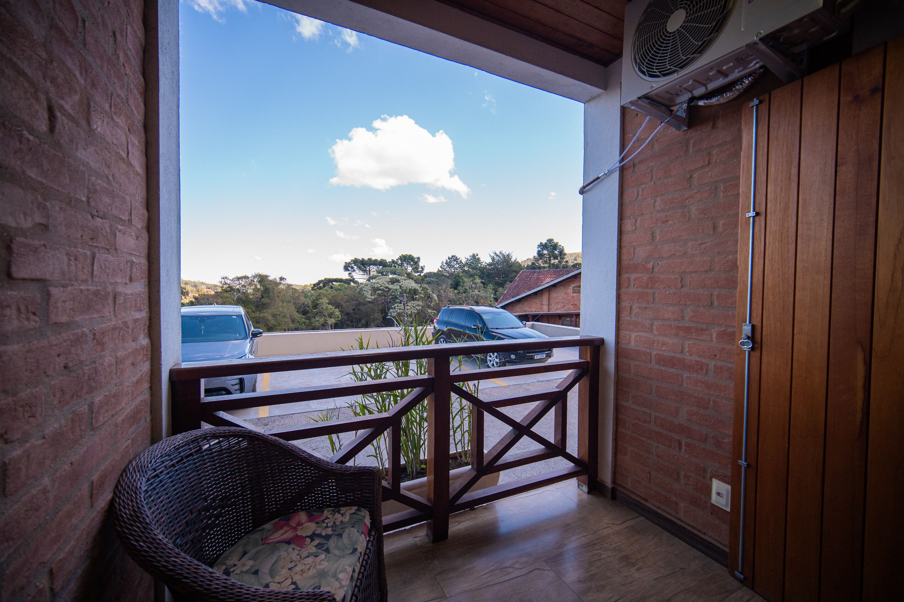 A balcony with a wicker chair, a view of parked cars, and a brick wall.