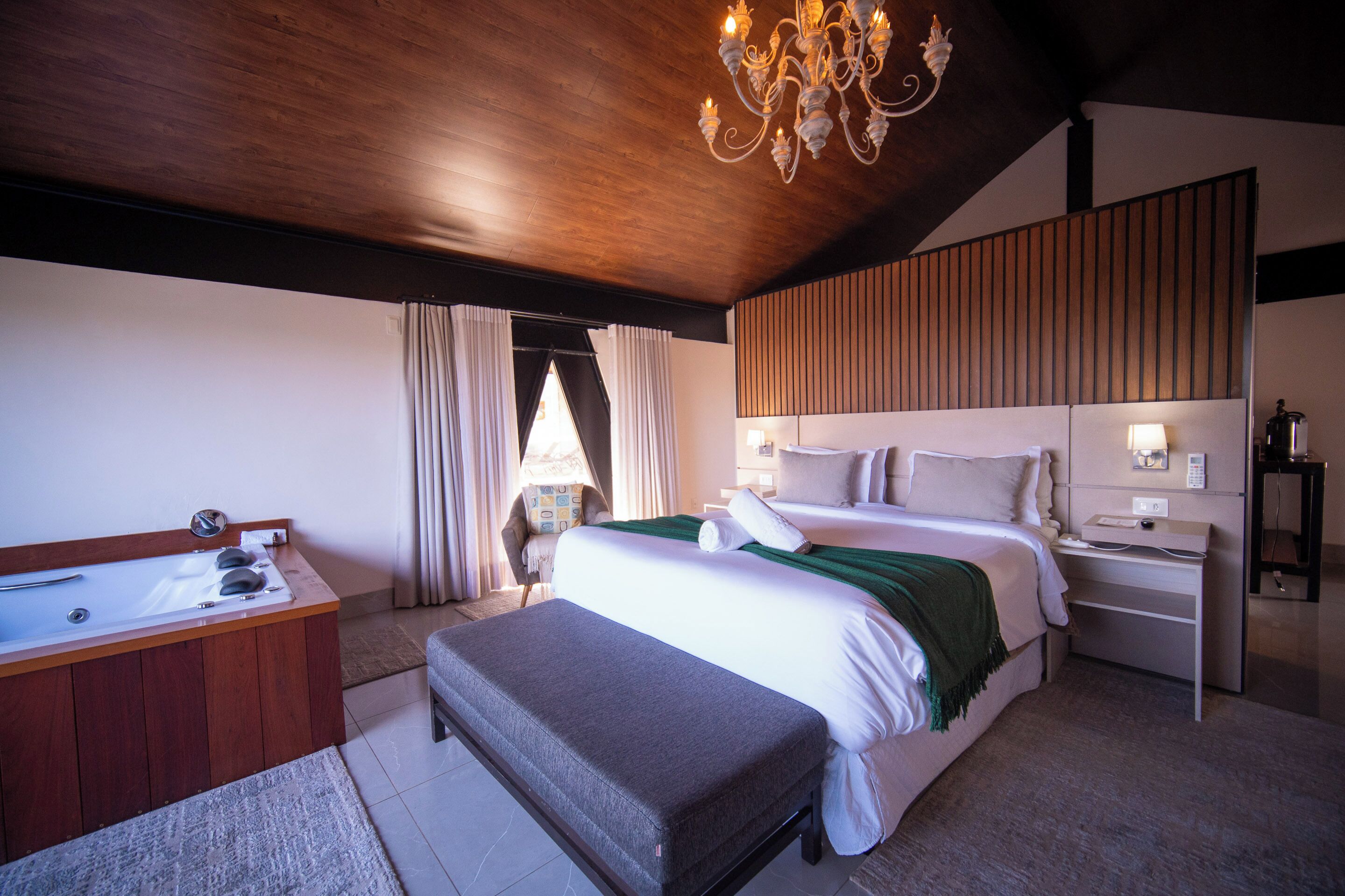 Ein Schlafzimmer mit einem großen Bett, einem Holzkopfteil, einem Nachttisch, einem Schreibtisch, einem Stuhl, einer Bank, einer Badewanne und einer Chandelier.
