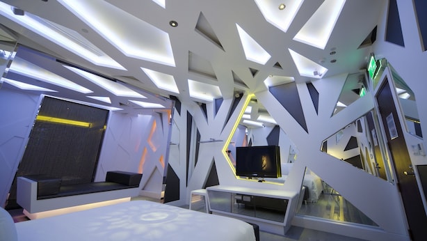 Quarto de hotel moderno com design geométrico no teto, televisão de tela plana e cama com cabeceira estampada.