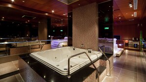 Uma área de spa moderna, com uma grande banheira de hidromassagem, mesa de massagem e espaço relaxante com sofá e televisão.