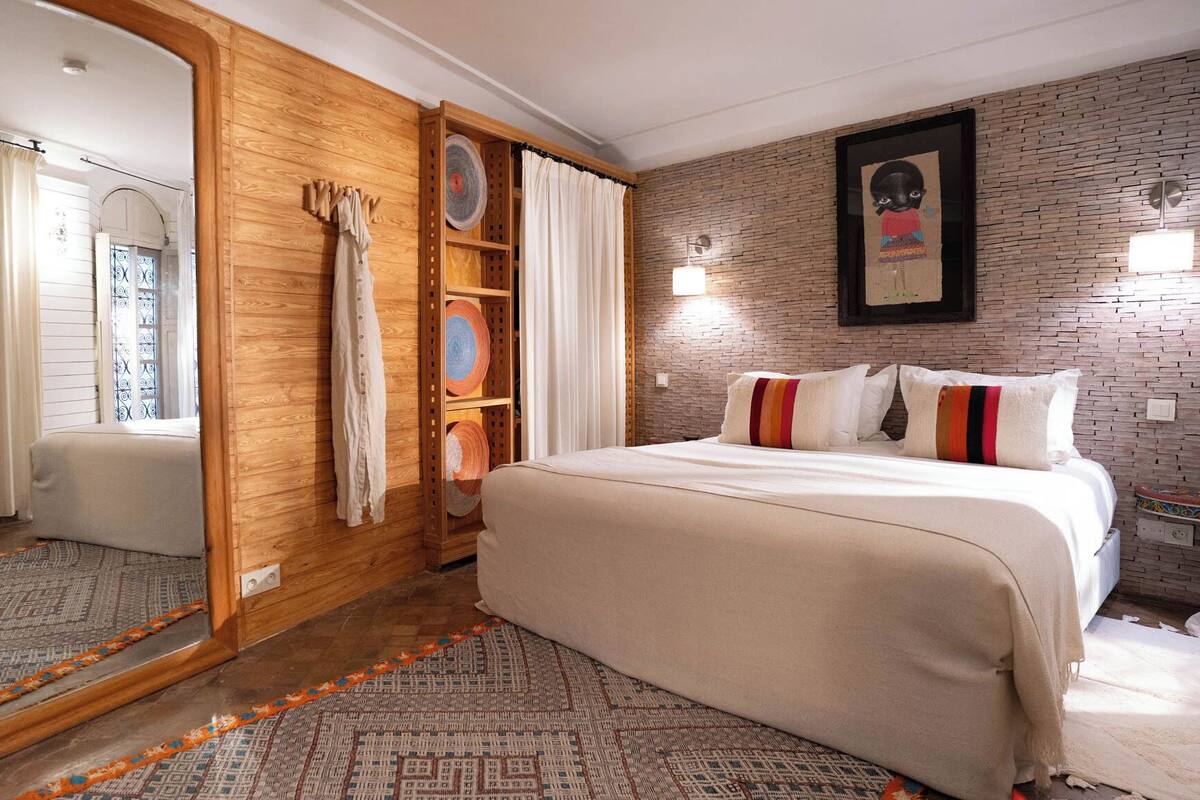 A bedroom with a large mirror, a bed with multiple pillows, a wooden wardrobe, and a brick-patterned wall.