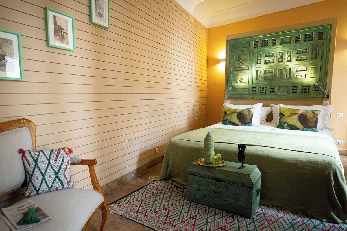 A bedroom with a wooden slat wall, a bed with a green headboard, a chair with a patterned cushion, and a small table with a bottle and glasses.