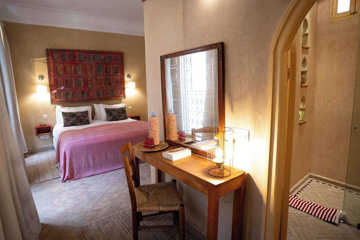 A hotel room with a bed, a desk with a chair, a mirror, and a window.