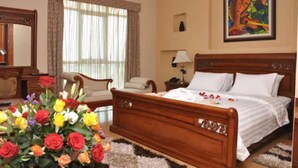 Habitación de hotel con una cama de madera, un ventanal con cortinas, un escritorio con lámpara y un cuadro en la pared.
