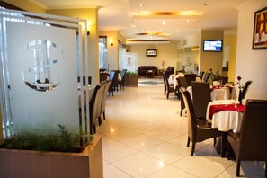 Un hall d’hôtel comprenant un comptoir d’accueil, un espace salon avec des tables et des chaises, un téléviseur fixé au mur et une plante décorative dans un coin.