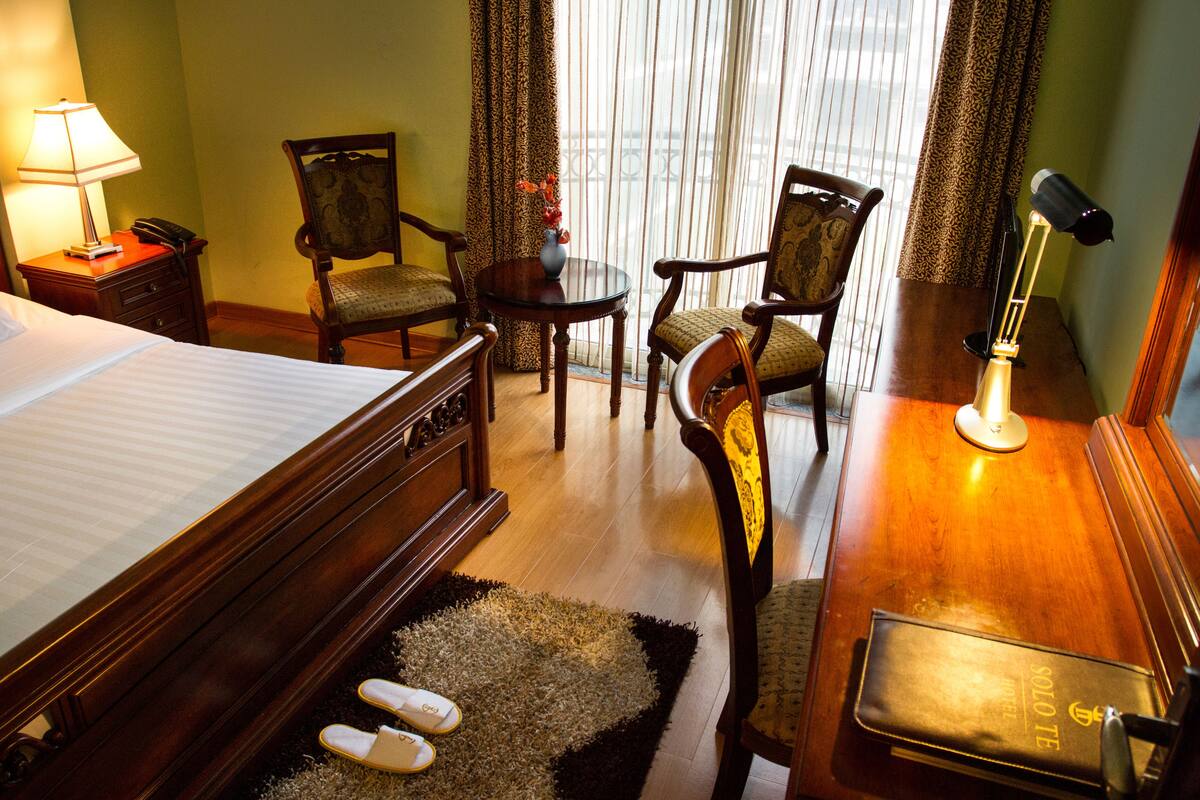 A hotel room with a bed, two chairs, a small table with a vase, a desk with a lamp, and a view through the window.