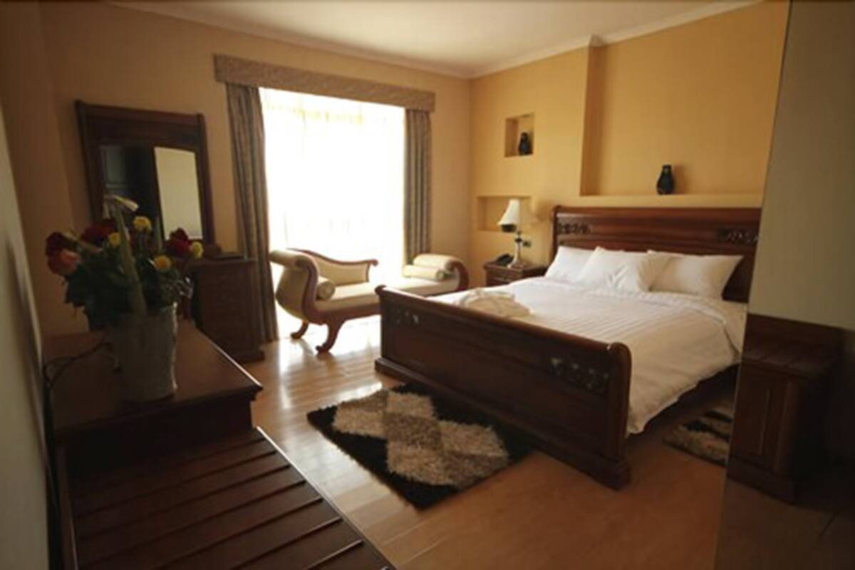 A hotel room with a wooden bed, a desk with a vase of flowers, a chair, a nightstand, and a window with curtains.