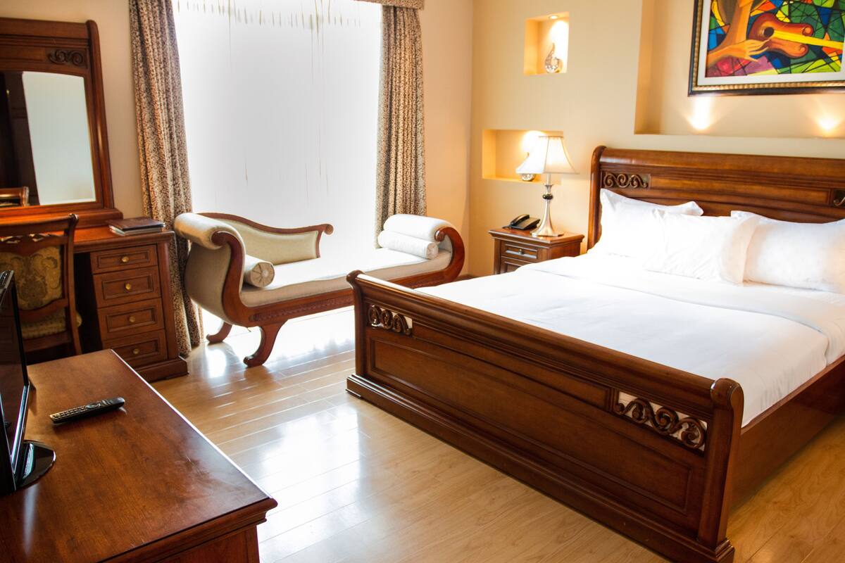 A hotel room with a wooden bed, a desk, a chair, and a nightstand with a lamp.