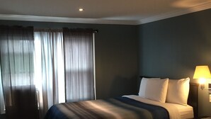 A hotel room with two beds, a nightstand, a lamp, and a window with curtains. - Jones Beach Hotel (Wantagh)