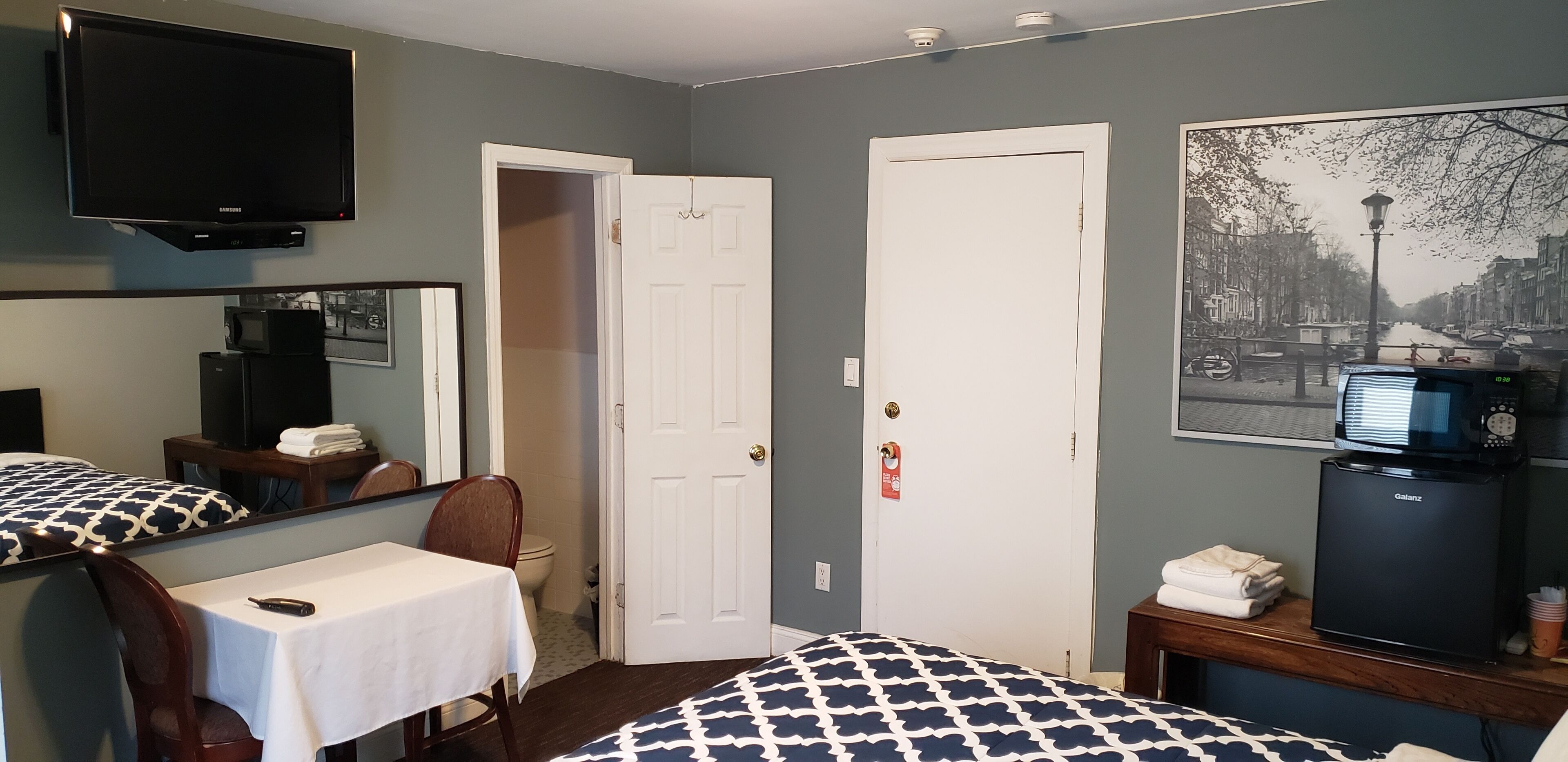 A hotel room with a bed, a desk with a chair, a television, a microwave, a refrigerator, and a door.