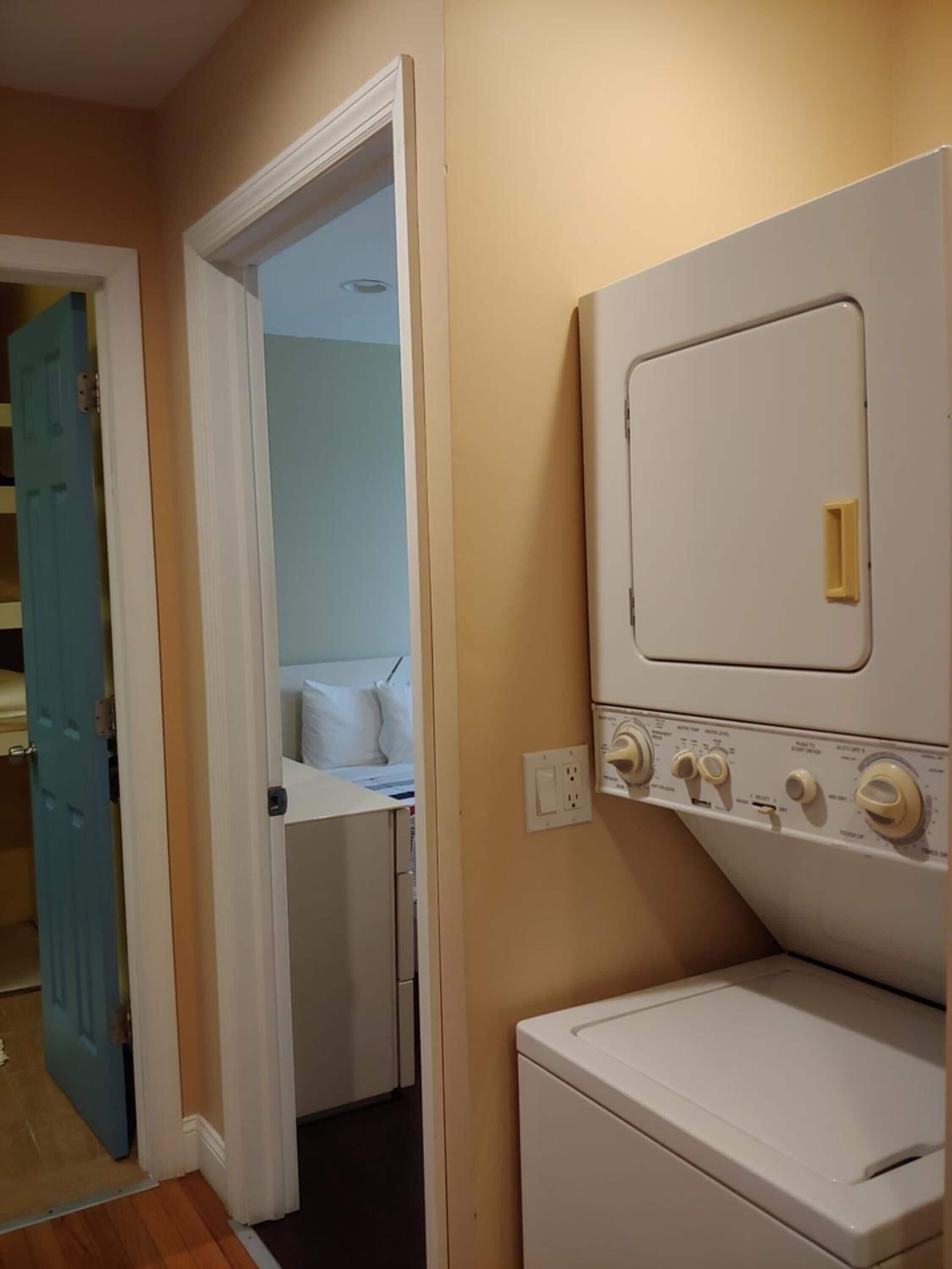 Une buanderie équipée d’un lave-linge et d’un sèche-linge à chargement frontal, une porte donnant sur une chambre et un interrupteur sur le mur.