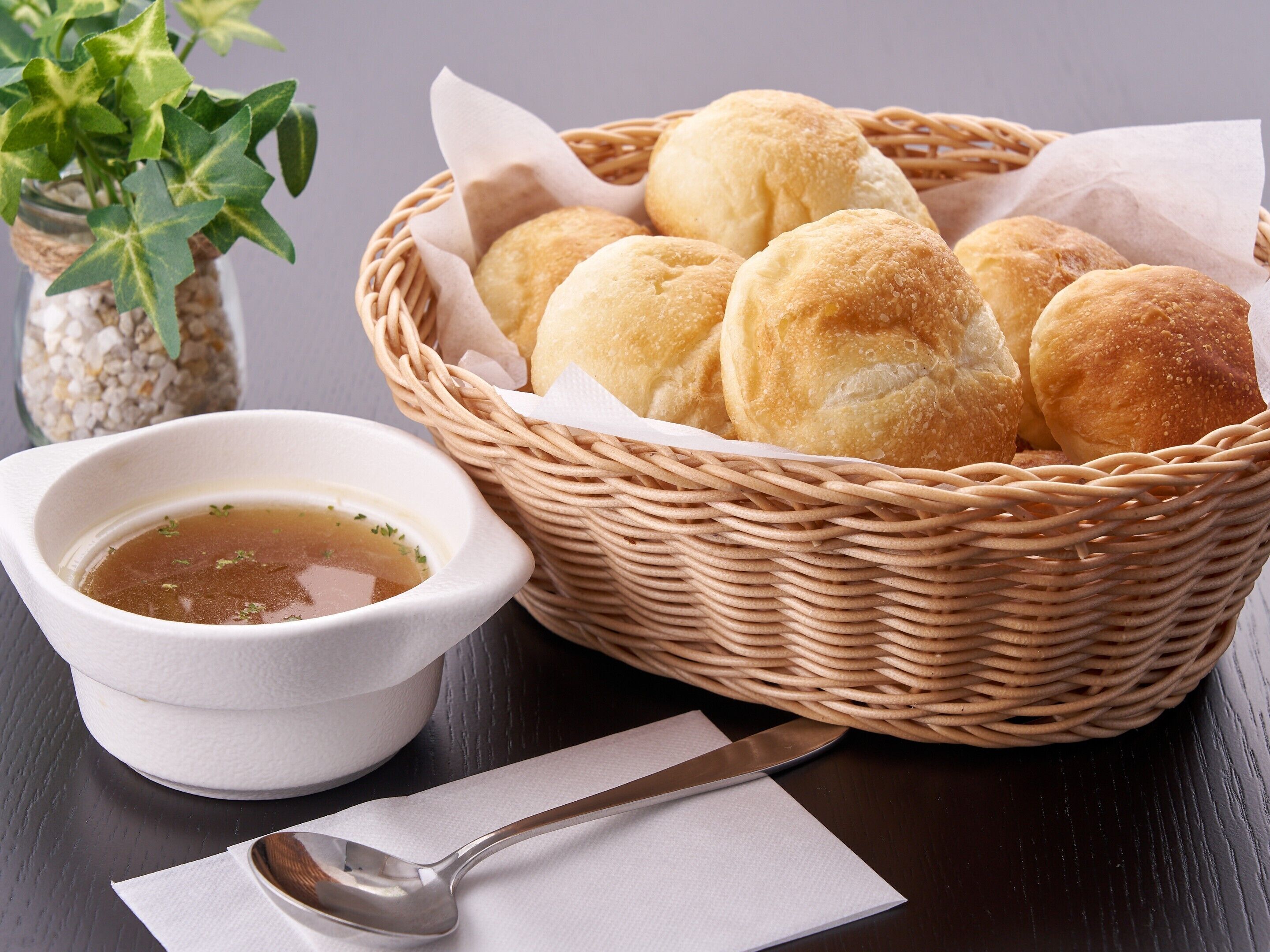A basket of round bread rolls with a dipping sauce, a spoon, and a napkin on a dark wooden table.