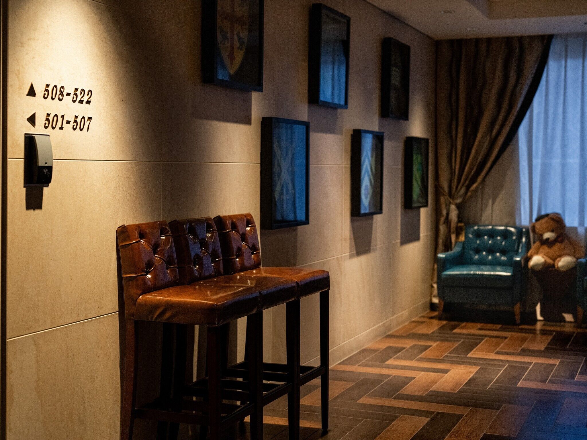 A hotel lobby with a leather bench, a blue sofa, and a teddy bear. There are framed pictures on the wall and a sign with phone numbers.