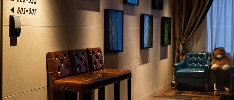 A hotel lobby with a leather bench, a blue sofa, and a teddy bear. There are framed pictures on the wall and a sign with phone numbers.