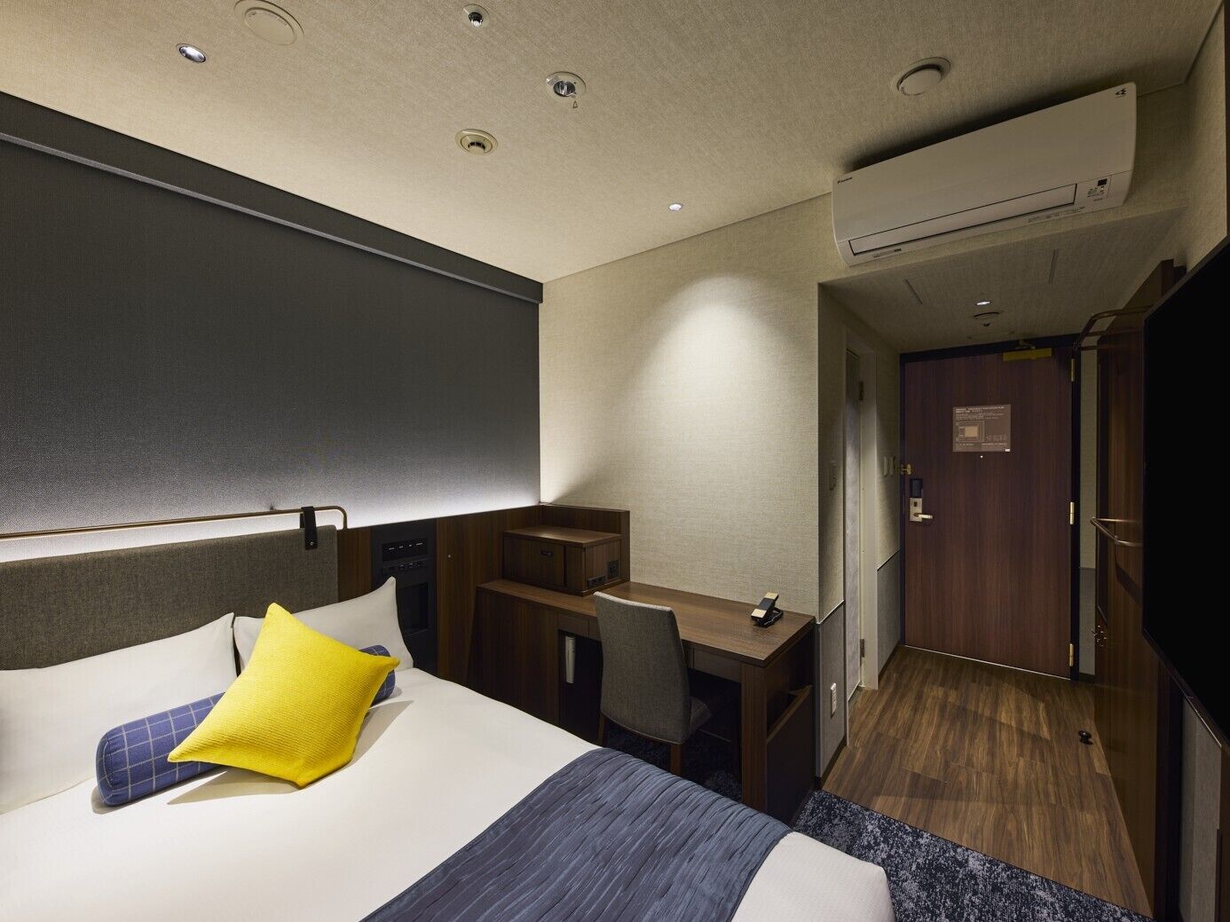 A hotel room with a bed, a desk, a chair, an air conditioning unit, and a door.