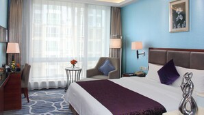Une chambre d’hôtel avec un grand lit, un bureau, une chaise, une petite table ornée de fleurs et une fenêtre avec des rideaux légers.