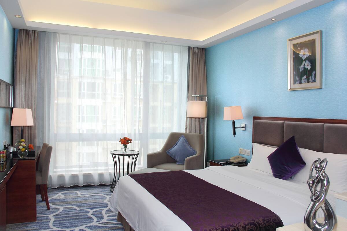 Une chambre d’hôtel avec un grand lit, un bureau, une chaise, une petite table ornée de fleurs et une fenêtre avec des rideaux légers.