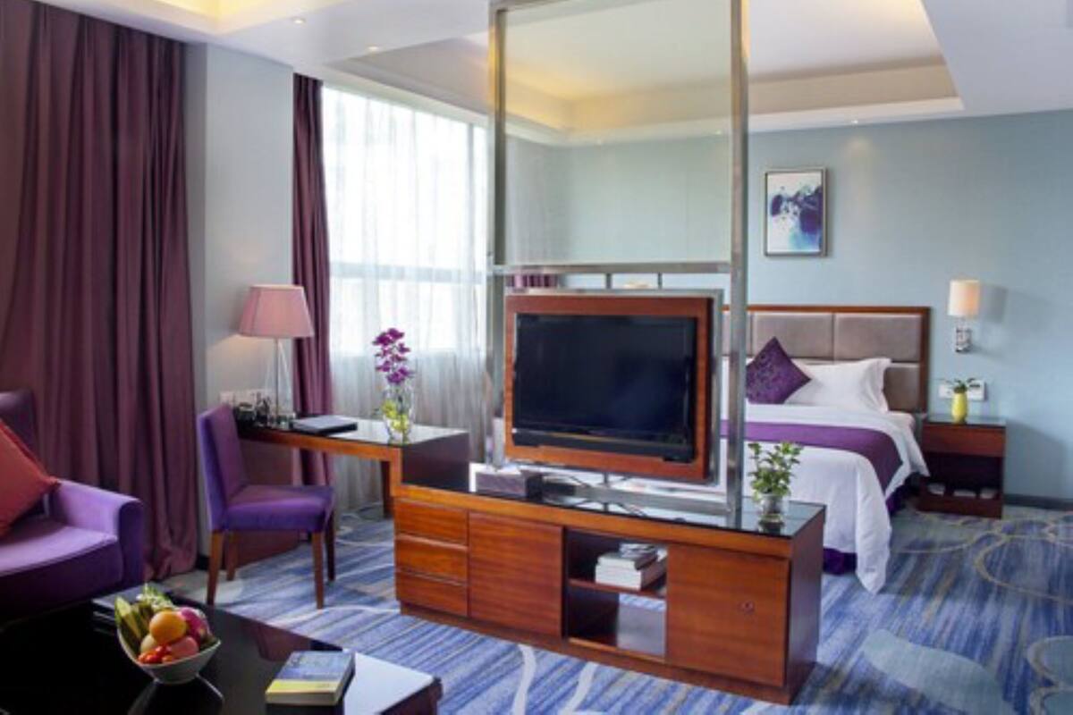 Une chambre d’hôtel dotée d’une grande télévision à écran plat, d’un bureau avec une lampe, d’un fauteuil violet, d’un lit avec une tête de lit et d’une moquette bleue.
