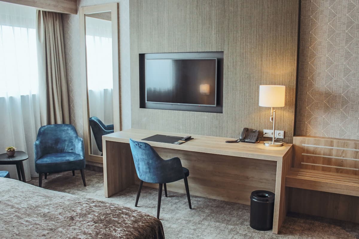 A hotel room with a wooden desk, two blue chairs, a TV mounted on the wall, a lamp, and a small trash bin.