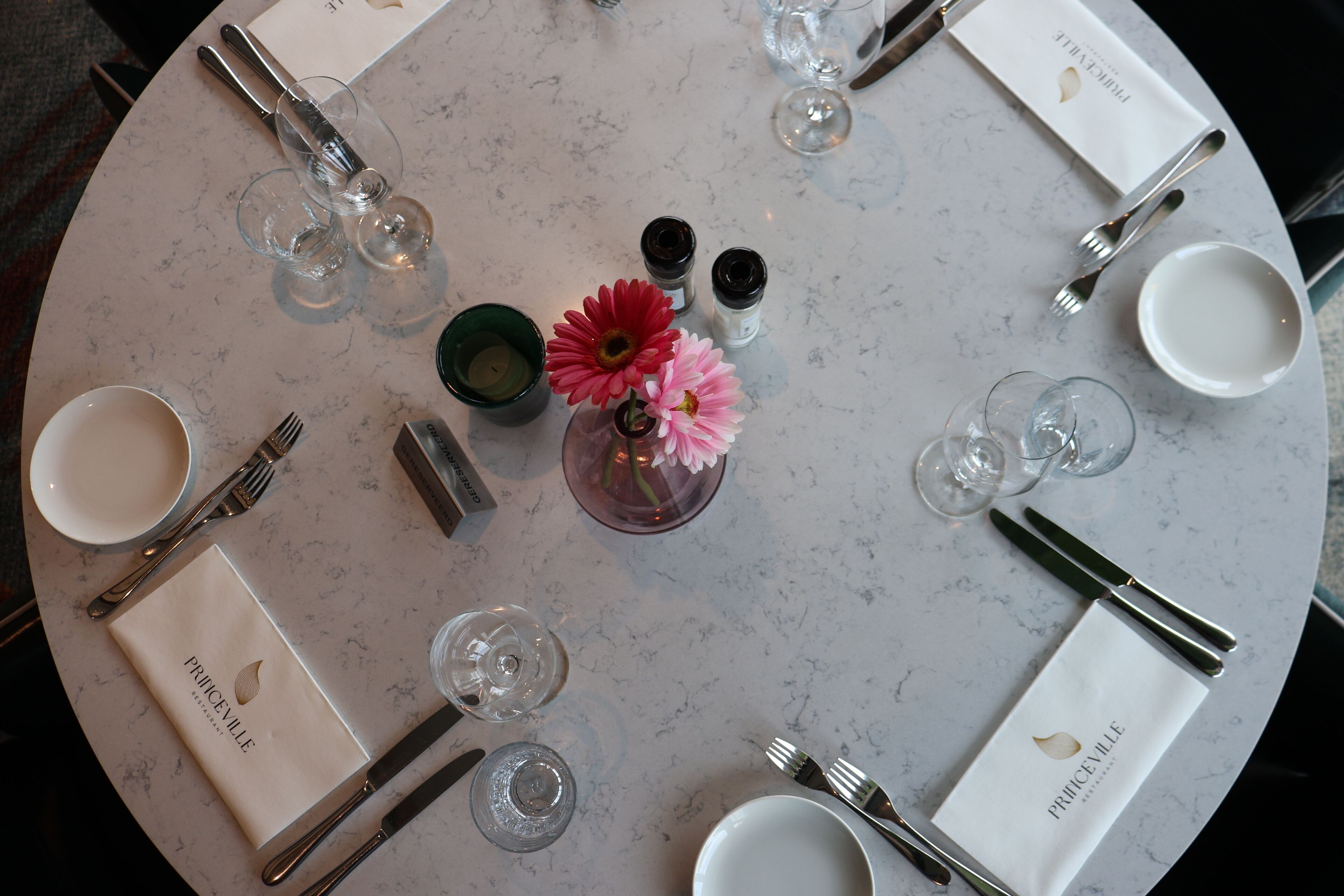 Una mesa redonda preparada para una comida, con un florero, saleros y pimenteros, y cristalería transparente.