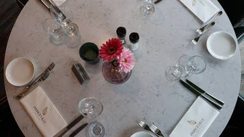 Una mesa redonda preparada para una comida, con un florero, saleros y pimenteros, y cristalería transparente.