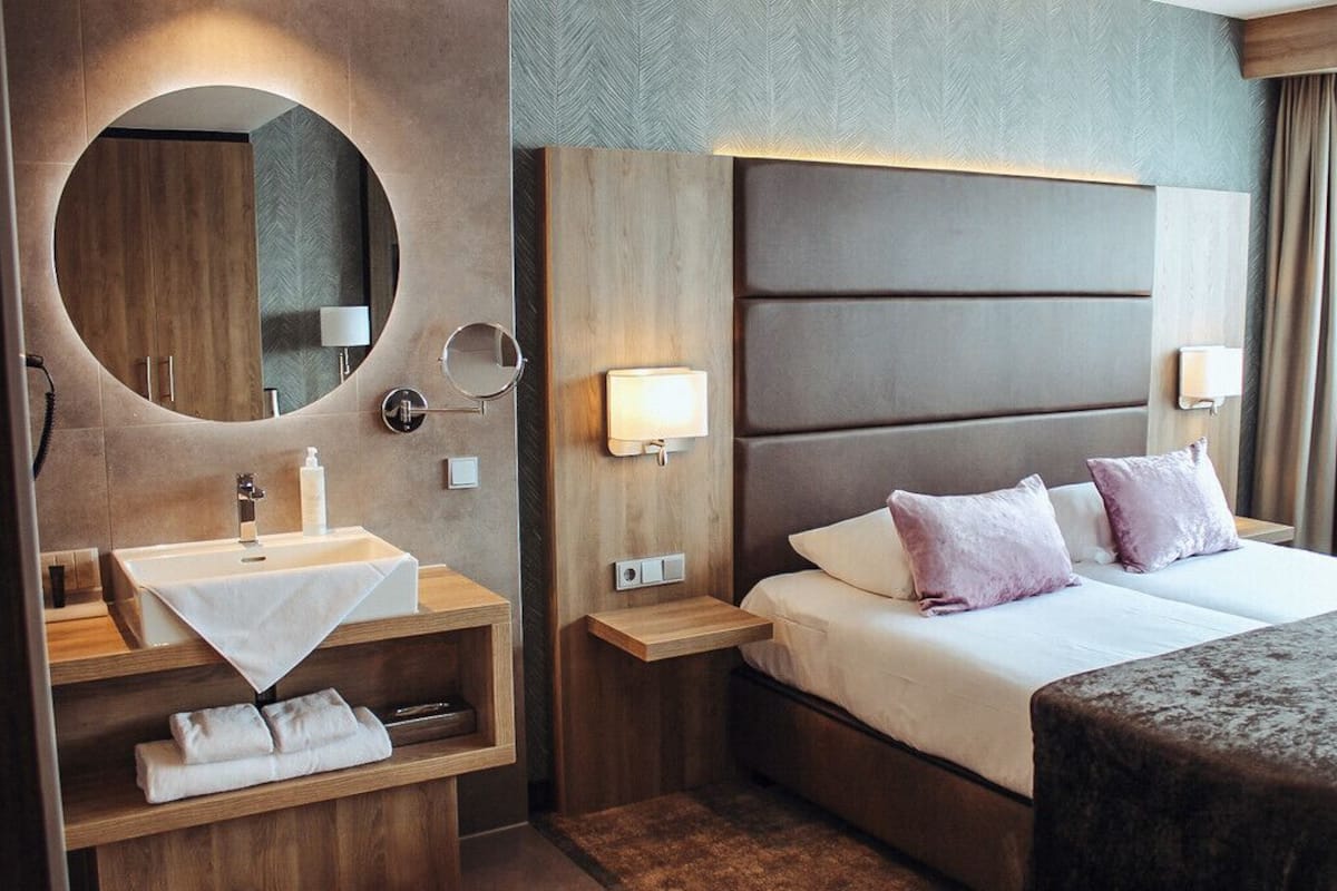 A modern hotel room with a large bed, a round mirror, a bedside table with a lamp, and a built-in sink.