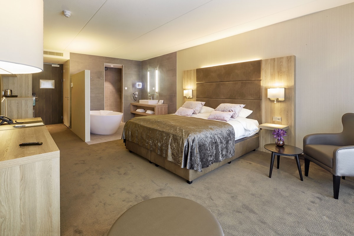 A hotel room with a large bed, a bedside table, a chair, a small table, a bathroom with a bathtub, and a mirror.