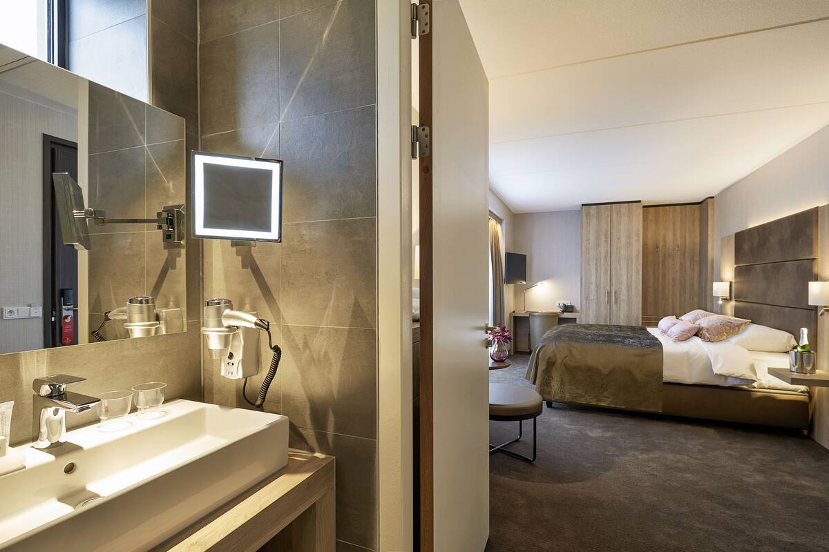 A modern hotel room with a large mirror, a sink with a vanity, a bed with a wooden headboard, and a bedside table with a lamp.