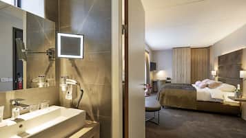 Et moderne hotelværelse med et stort spejl, en vask med tilhørende underskab, en seng med trægavl og et natbord med lampe.