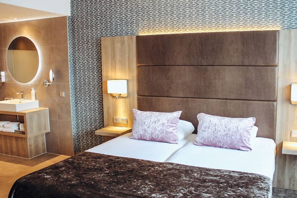 A hotel room with a large bed, two bedside lamps, a wooden headboard, a built-in shelf with towels, and a patterned wall.