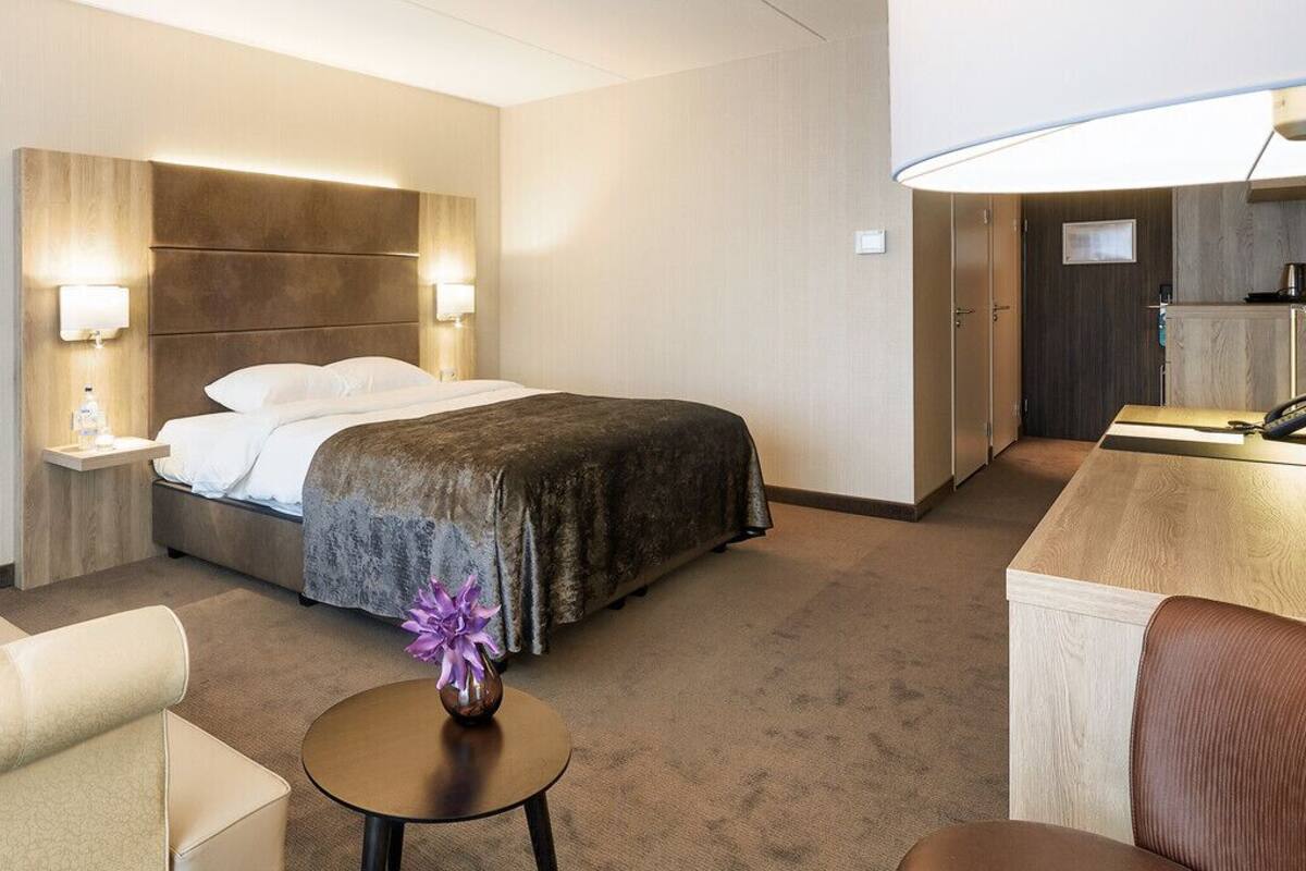 A hotel room with a bed, bedside tables, a desk, a chair, and a small table with a vase.