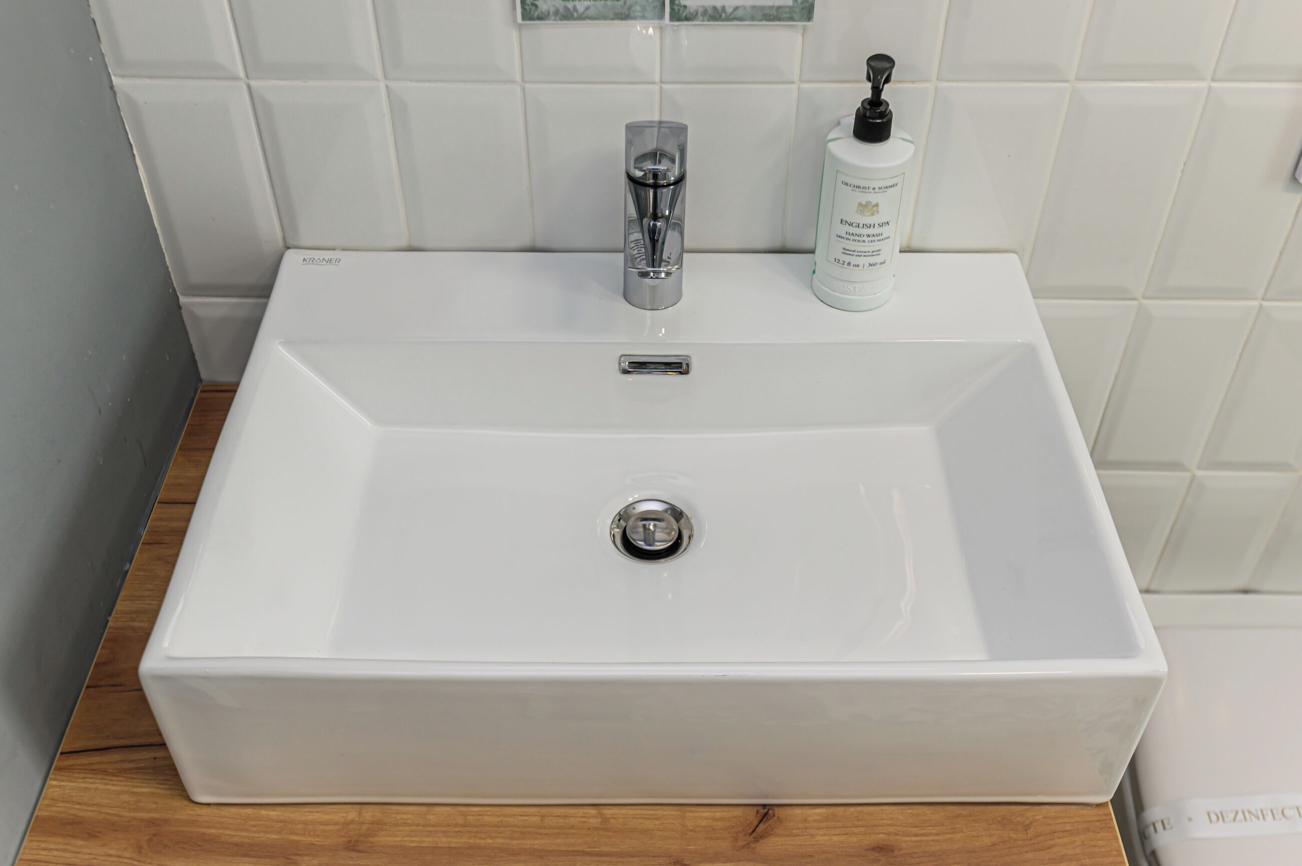 Uma pia de banheiro retangular branca com uma torneira moderna, um frasco de sabonete líquido e uma pequena garrafa de shampoo.