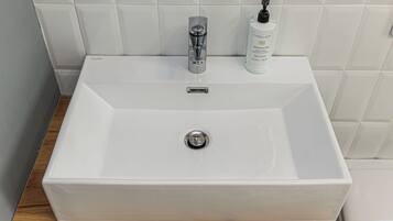 Uma pia de banheiro retangular branca com uma torneira moderna, um frasco de sabonete líquido e uma pequena garrafa de shampoo.