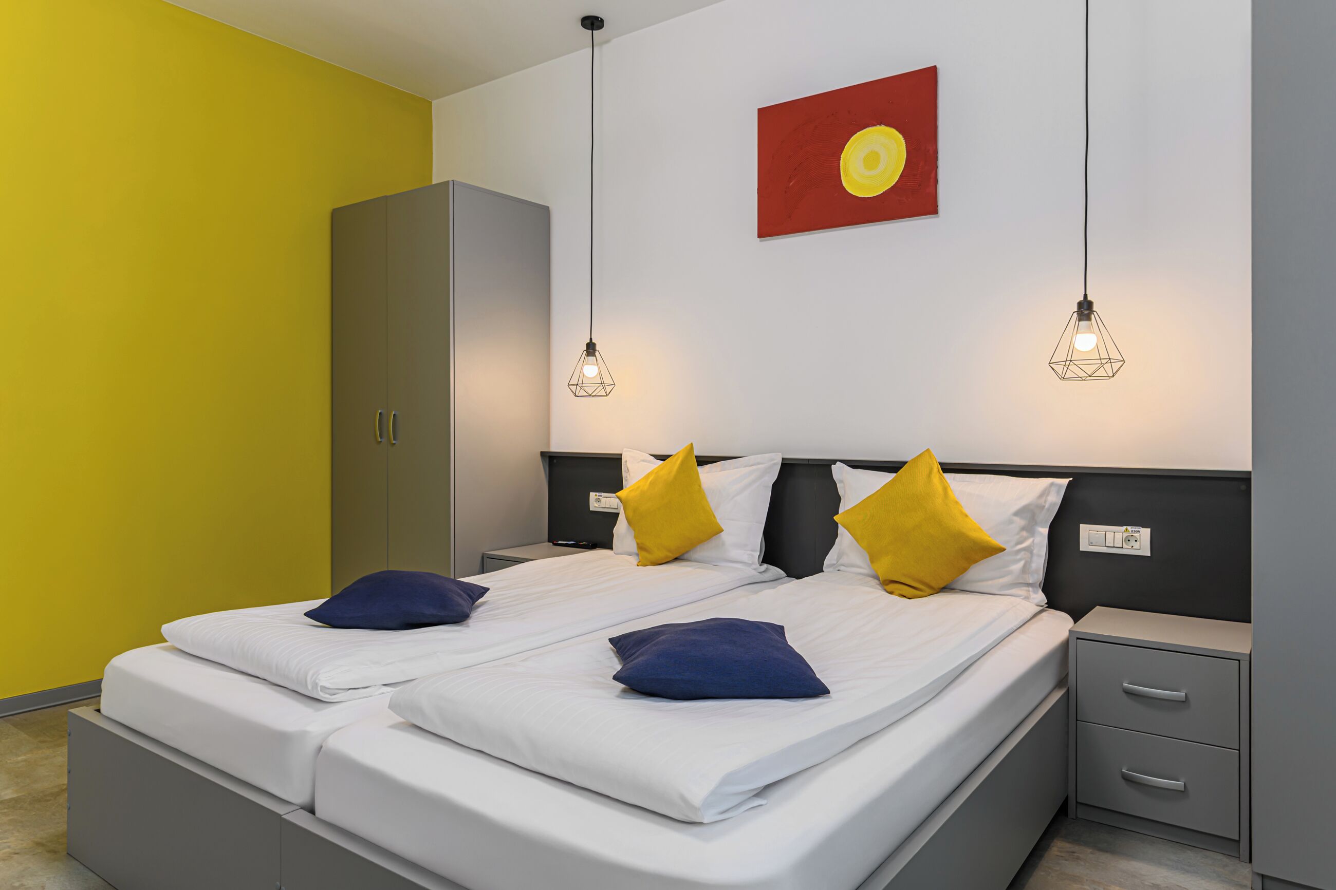 A double bed with white linens, two yellow and two blue pillows, a grey wardrobe, and a red and yellow wall decoration.