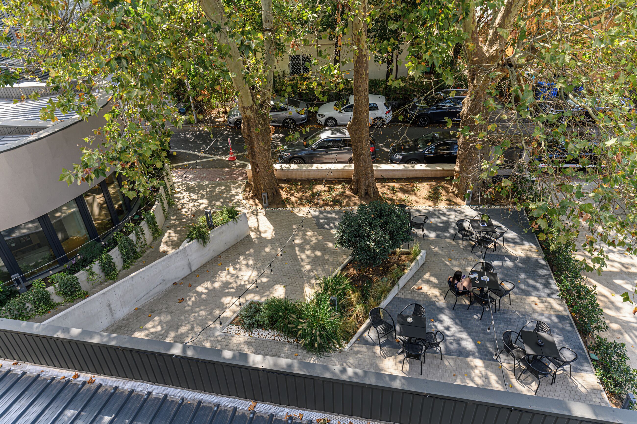 Un espacio al aire libre con mesas y sillas, rodeado de árboles y un estacionamiento con coches.