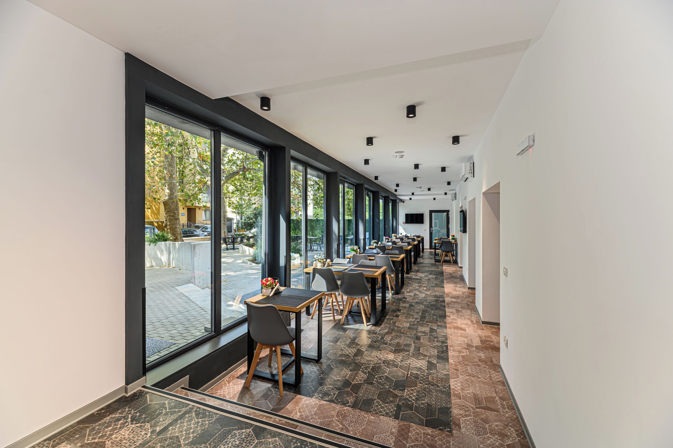 Un pasillo moderno de hotel con ventanas amplias, piso de baldosas y una fila de mesas y sillas.