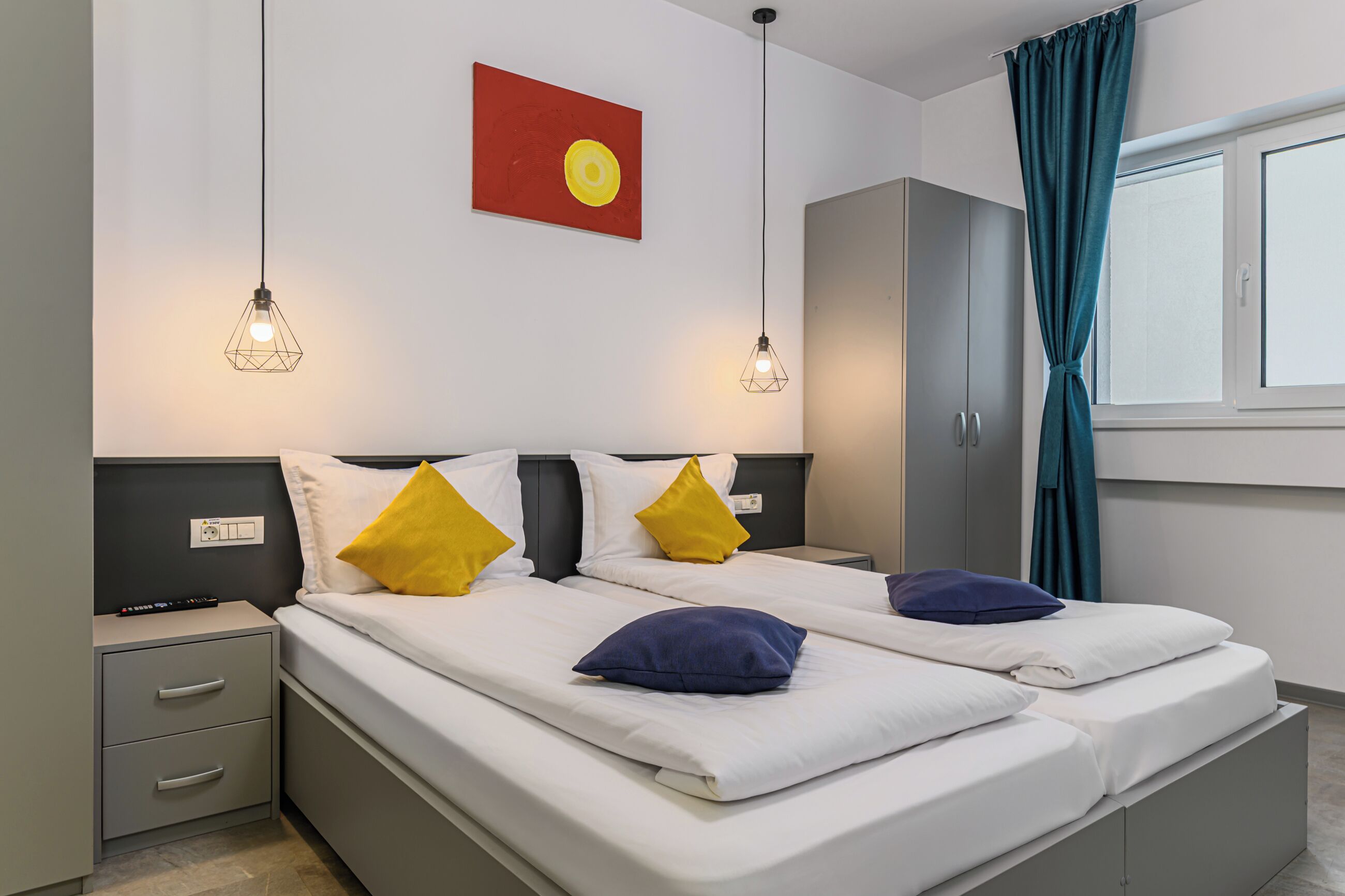 A double bed with white bedding and two yellow pillows, a nightstand with a remote control, and a red abstract painting on the wall.